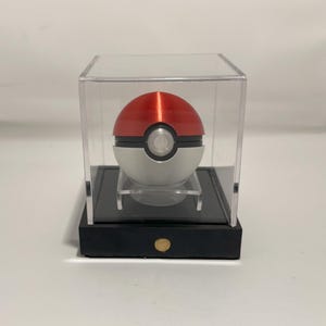 May include: A red and white Poké Ball replica, presented in a clear acrylic display case. The ball features a silver central section and sits on a black base with a gold-coloured button. The case is designed to showcase the collectible item.