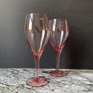 May include: Two pink-tinted wine glasses with long stems and round bases. The glasses are made of clear glass and stand on a marble-patterned surface against a dark background. The glasses are empty.