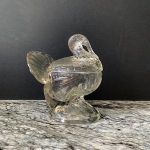 May include: A clear glass turkey-shaped candy dish with a lid. The dish has a detailed textured design and sits on a pedestal base. The turkey's tail feathers and head are clearly defined. The background is dark.