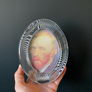 May include: Oval glass picture frame with a portrait of Vincent van Gogh. The frame has a wavy, textured design. The portrait features the artist with a red beard and a purple jacket.