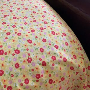 May include: A yellow fabric featuring a dense, repeating floral pattern. The design consists of small, five-petaled flowers in vibrant red, soft pink, and pale orange, interspersed with tiny white and green circular accents across the surface.