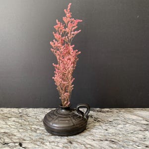 May include: A small, dark gray ceramic vase with a handle, holding a bundle of dried pink flowers. The vase has a geometric pattern around its body. The flowers are a light pink color and add a touch of color to the scene.
