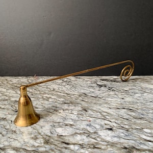 May include: A brass candle snuffer with a bell-shaped base and a long, straight handle ending in a spiral design. The snuffer is made of shiny, golden-colored metal and sits on a marble-patterned surface.