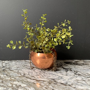 May include: A round, metallic copper-colored planter with a smooth, reflective surface. The planter is suspended by thin, matching copper-toned metal chains attached to its sides. A lush, green artificial plant with small, rounded leaves fills the pot.