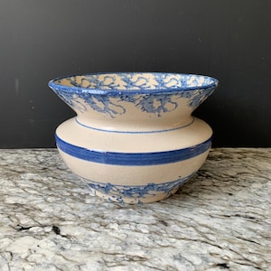 May include: A ceramic vase with a wide, flared rim and a bulbous body. The vase is cream-colored with blue abstract patterns around the rim and a horizontal band around the body. The vase is likely handmade.