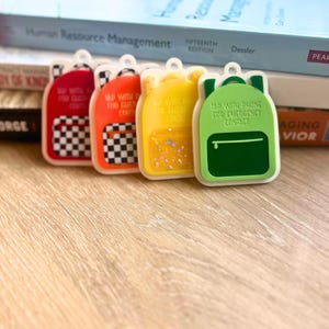 May include: Four colorful backpack-shaped keychains in red, orange, yellow, and green. Each keychain has a white border and a small loop at the top. The green keychain reads "TAP WITH PHONE FOR EMERGENCY CONTACT".