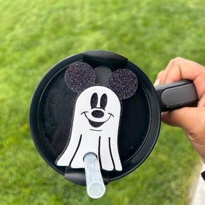 May include: A black and white ghost with Mickey Mouse ears and a smiling face. The ghost is attached to a black lid with a straw.