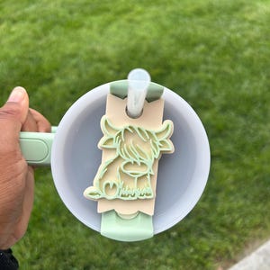 May include: A green and white Highland cow design on a beige background. The design is attached to a green and white lid for a reusable water bottle.