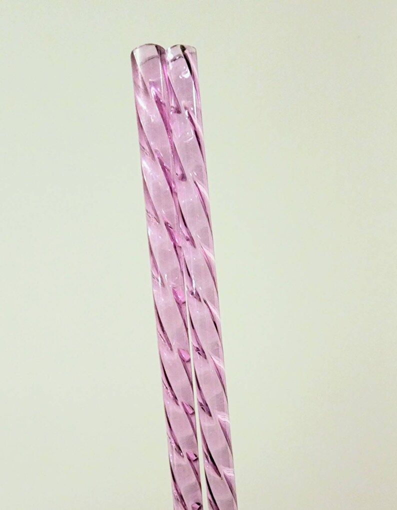 Pu&ograve; includere: Due cannucce da bere in vetro rosa con un design intrecciato.