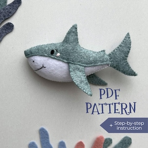 May include: A felt shark toy in shades of green and white, with a smiling face and stitched details. The image includes the text "PDF PATTERN" and "Step-by-step instruction". Decorative felt coral accents are in the corners.
