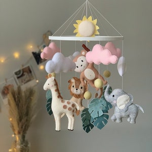 May include: A handmade baby mobile featuring felt safari animals: a giraffe, lion, monkey, elephant, and sun. The mobile includes pink and white clouds, green leaves, and yellow accents. The mobile is suspended from a white frame.