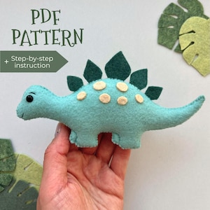 May include: A teal felt dinosaur toy with dark green spikes and tan spots. The image includes the text "PDF PATTERN" and "Step-by-step instruction". Decorative felt leaves are also visible.