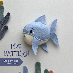 Könnte beinhalten: Ein hellblauer Filz-Hai mit weißem Bauch, gestickten Details und einem lächelnden Gesicht. Das Bild enthält den Text "PDF PATTERN" und "Step-by-step instruction". Dekorative Filzkorallen in Blau-, Grün- und Rosatönen sind ebenfalls vorhanden.