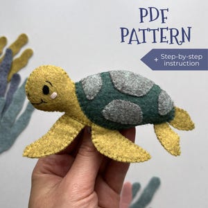 May include: A handmade felt turtle in shades of yellow, green, and grey. The turtle has a detailed design with a dark green shell and grey spots. The image includes the text "PDF PATTERN + Step-by-step instruction".
