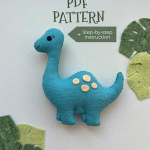 May include: A turquoise felt dinosaur with tan spots and black eyes. The image includes the text "PDF PATTERN" and "Step-by-step instruction". Green felt leaf cutouts are scattered around the dinosaur.