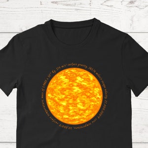 May include: Black t-shirt with a graphic of the sun. The graphic includes text about the sun's temperature, mass, surface gravity, and escape velocity.