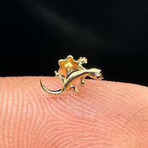 Può includere: Un minuscolo ciondolo a forma di lucertola dorata con dettagli intricati. La lucertola è raffigurata con la coda arricciata e le zampe distese. Il ciondolo è mostrato sulla punta di un dito su sfondo nero. Il design della lucertola include un volant dietro la testa.