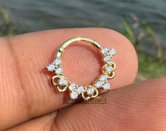 May include: A gold-colored septum ring with a floral design. The ring is adorned with small, clear gemstones. The jewelry is held in a person's hand, with a blurred background of water and greenery.