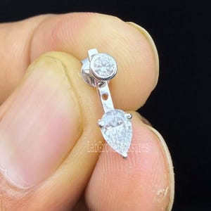 May include: A silver-toned earring jacket featuring a small round brilliant-cut stone at the top and a larger pear-shaped stone suspended below by a vertical metal bar. The piece has a polished metallic finish and clear faceted stones.