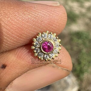Pode incluir: Um anel de nariz de ouro com uma gema rosa cercada por gemas brancas menores. O anel é segurado por um dedo.