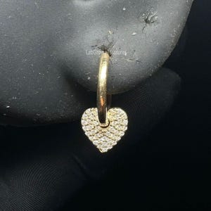 May include: A gold hoop earring with a heart-shaped charm. The heart is encrusted with small, sparkling stones. The earring is shown on a black background.