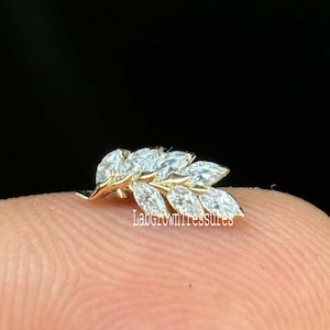 May include: A delicate gold-toned leaf-shaped earring with marquise-cut gemstones. The earring features a cluster of sparkling stones arranged to resemble leaves. The image is set against a dark background, highlighting the jewelry's intricate design.