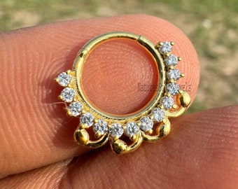 May include: A gold-colored septum ring with a decorative design. The ring features a series of small, clear gemstones set around the outer edge. The design is circular with a floral-like pattern.