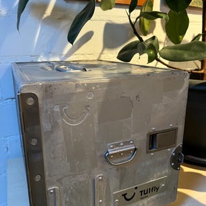Könnte beinhalten: Ein rechteckiger, silberfarbener Metallkoffer mit Gebrauchsspuren. Er hat einen Griff, einen Verschluss und den Text "TUIfly Nordic" auf einem kleinen Schild. Der Koffer steht auf einer hellen Oberfläche, mit sichtbaren Nieten und Nähten.