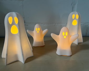 Könnte beinhalten: Vier weiße, geisterförmige Dekorationslichter mit leuchtend gelben Augen und Münder. Die Lichter sind auf einer Holzoberfläche vor einer weißen Backsteinmauer angeordnet. Die Lichter variieren in der Größe und erzeugen ein gruseliges Halloween-Thema.