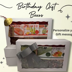 May include: Three white gift boxes with clear windows showing different flavored dip mixes. The boxes are stacked on top of each other. The top box has a red, green, and yellow fiesta dip mix. The middle box has a green spinach dip mix and a green chive dip mix. The bottom box has a green vegetable dip mix. The boxes are decorated with a gray bow. The text "Birthday Gift Boxes" is written in black cursive above the boxes. The text "Personalize your Gift message" is written in black text to the right of the boxes.