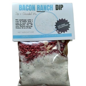 May include: A small white bag with a clear front containing a mix of red, white, and beige ingredients. The bag has a label that reads "Bacon Ranch Dip, Dip & Cheeseball Mix, Fat Free Sugar Free, Net Weight 1 oz, Dipdelivered.com for recipes & more".