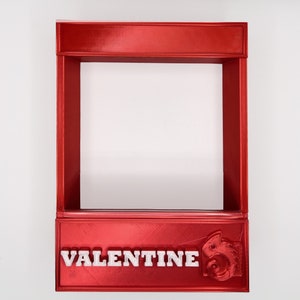 May include: A red 3D printed frame with the word "VALENTINE" printed in white on the bottom. The frame is empty and has a small, white, 3D printed chameleon on the right side.