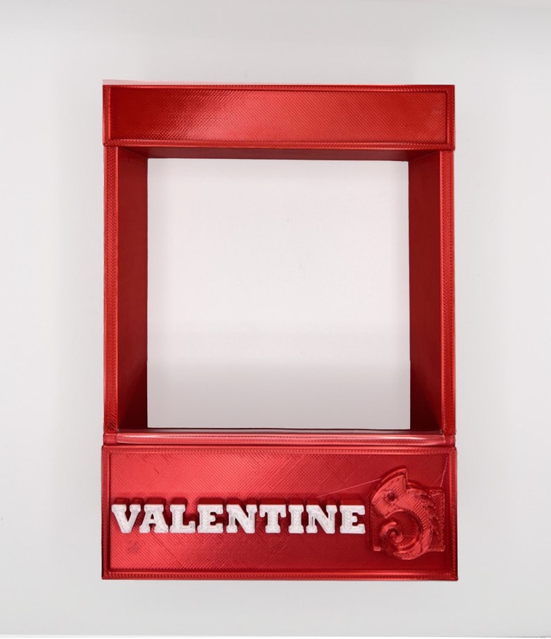 May include: A red 3D printed frame with the word "VALENTINE" printed in white on the bottom. The frame is empty and has a small, white, 3D printed chameleon on the right side.