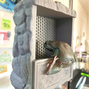 Puede incluir: Un hábitat para reptiles impreso en 3D de color gris con un diseño de roca texturizado. Una rana arborícola blanca está posada en el borde del hábitat.