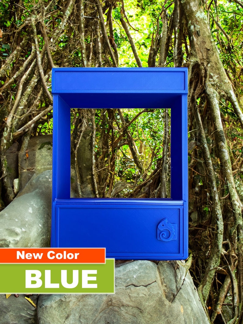 May include: A blue, rectangular, open-top display case with a small chameleon logo on the front. The case is sitting on a rock in a forest setting.