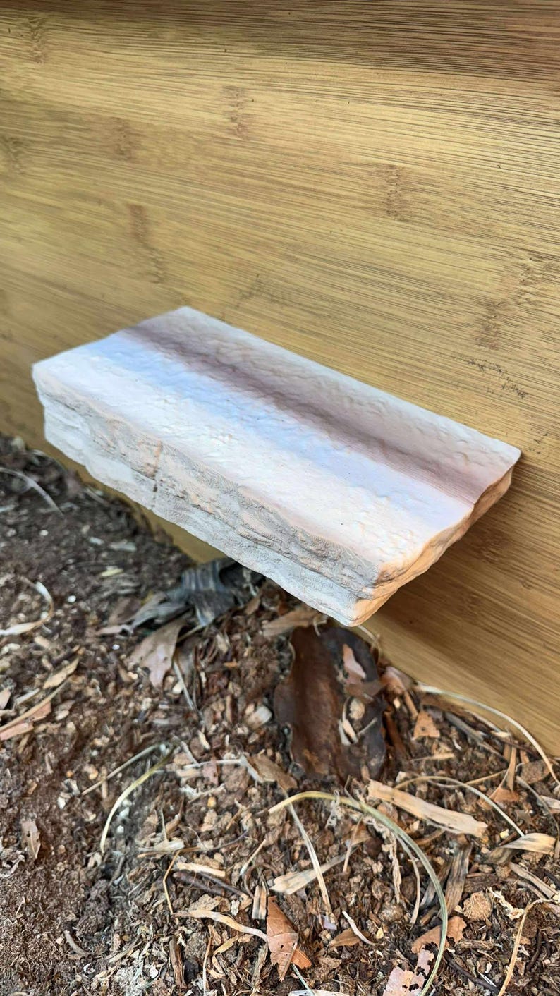 K&ouml;nnte beinhalten: Ein rechteckiges Regal aus Naturstein mit einem hellrosa und braunen Farbverlauf. Das Regal wird von einer Holzkonstruktion getragen und ruht &uuml;ber einem Bett aus Erde und Bl&auml;ttern. Der Stein hat eine raue, strukturierte Oberfl&auml;che.