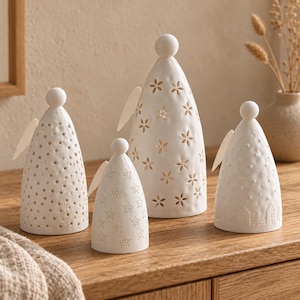May include: A set of four white ceramic cone-shaped figurines with spherical heads and small wings. Each piece features unique cut-out patterns including stars, flowers, and a landscape with trees and houses, allowing light to pass through.