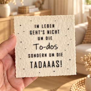 Può includere: Una targa quadrata color bianco sporco con una superficie testurizzata e scritte nere in rilievo che recitano: IM LEBEN GEHT'S NICHT UM DIE To-dos SONDERN UM DIE TADAAAS! La superficie ha un motivo organico irregolare e piccoli punti neri.
