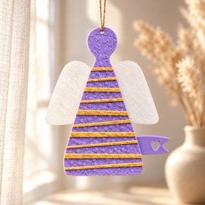 Puede incluir: Un adorno colgante con forma de ángel de color morado con una superficie texturizada y rayas doradas horizontales. La figura presenta dos alas blancas translúcidas y una pequeña bandera morada con un recorte en forma de corazón al final.