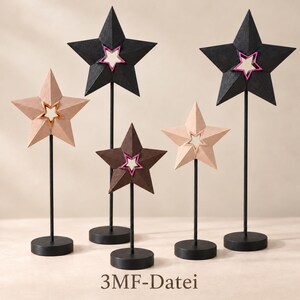 May include: Five star-shaped ornaments on thin black metal rods with circular bases. Two stars are black with pink centers, one is dark brown with a pink center, and two are light beige with gold centers. The text 3MF-Datei is visible.