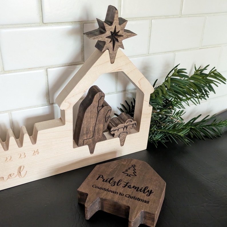 May include: Wooden Christmas nativity scene with a star on top. The scene includes figures of Mary, Joseph, and baby Jesus. A separate wooden piece reads "Pritzl Family Countdown to Christmas."