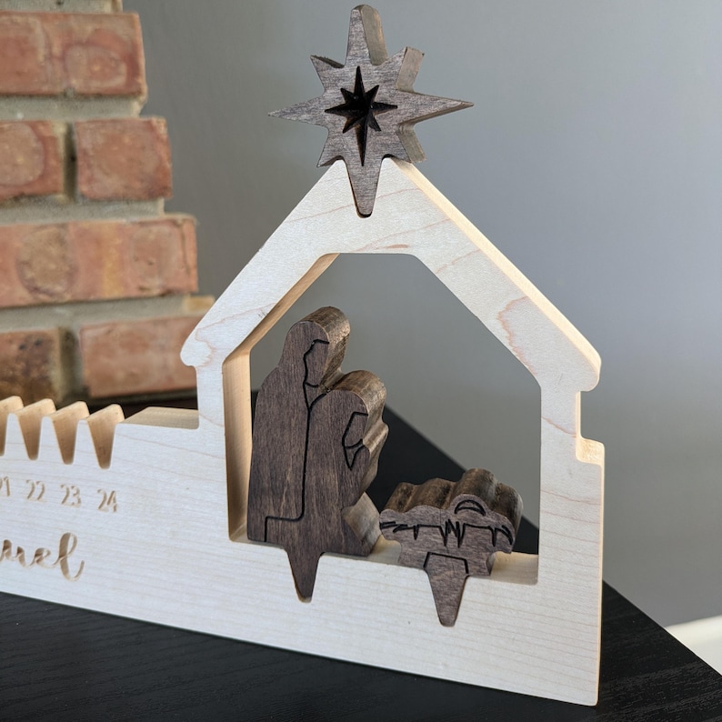 May include: Wooden nativity scene with a star on top. The scene features a house-shaped structure with figures inside. The star and figures are a darker wood, contrasting with the lighter wood of the house. The numbers 21, 22, 23, and 24 are visible.