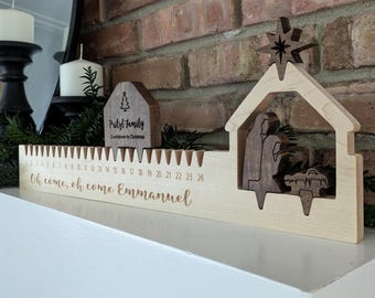 Könnte beinhalten: Weihnachts-Adventskalender aus Holz mit einer Krippenszene und einem Stern. Der Kalender hat eine Zählung von 1 bis 24, mit den Worten "Oh come, oh come Emmanuel" eingraviert. Ein kleines Holzhaus mit dem Text "Pritzl Family" ist ebenfalls vorhanden.