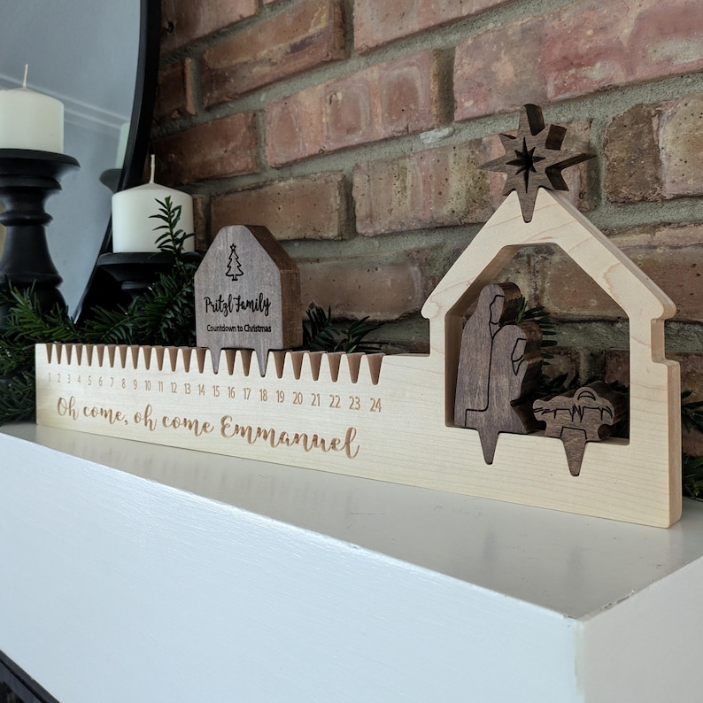 May include: Wooden Christmas advent calendar with a nativity scene and star topper. The calendar has a countdown from 1 to 24, with the words "Oh come, oh come Emmanuel" engraved on the base. A small wooden house with the text "Pritzl Family" is also present.