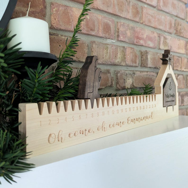 May include: A wooden Christmas advent calendar with a light wood finish. The calendar has 24 numbered slots and the words "Oh come, oh come Emmanuel" are engraved on the front. Includes wooden figurines and a small house-shaped decoration.