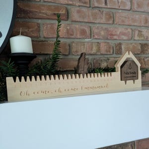 May include: A wooden Christmas countdown calendar with a house-shaped design and the text "Pristl Family Countdown to Christmas." The calendar has a ruler-like design with numbers 1-24 and the words "Oh come, oh come Emmanuel."