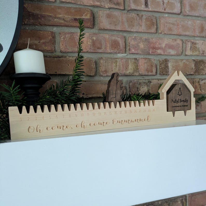 May include: A wooden Christmas countdown calendar with a house-shaped design and the text "Pristl Family Countdown to Christmas." The calendar has a ruler-like design with numbers 1-24 and the words "Oh come, oh come Emmanuel."