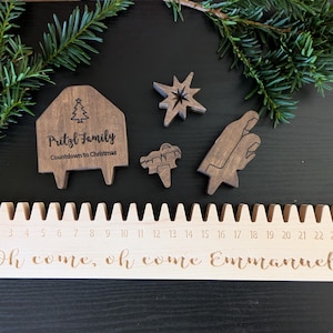 May include: Wooden Christmas countdown calendar with laser-cut details. Includes a house-shaped piece with "Pritzl Family" and "Countdown to Christmas" text, plus star, manger, and other shapes. The calendar has numbers 3-22 and the phrase "Oh come, oh come Emmanuel".