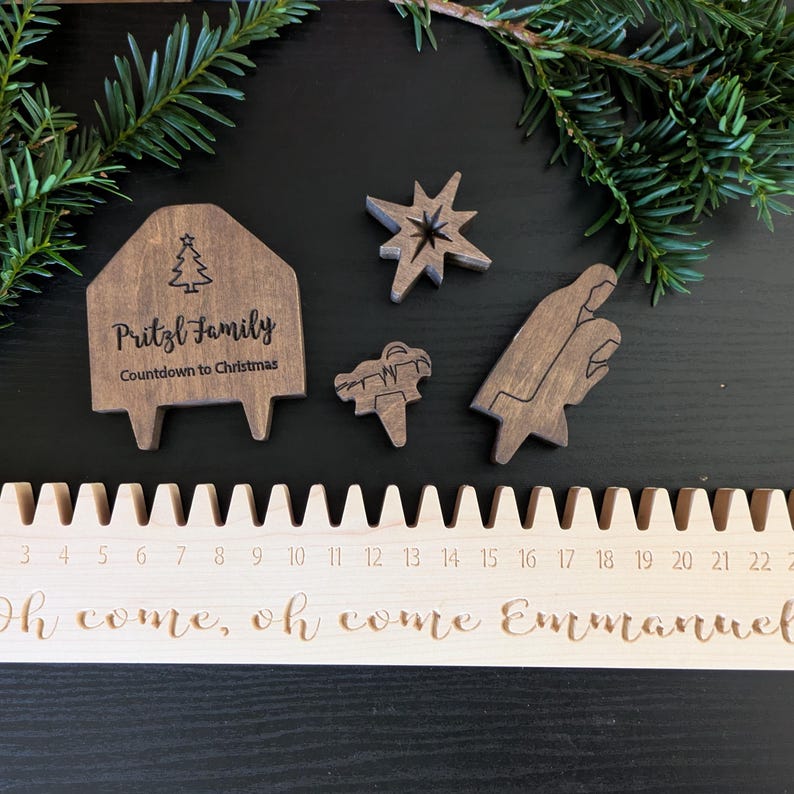 May include: Wooden Christmas countdown calendar with laser-cut details. Includes a house-shaped piece with "Pritzl Family" and "Countdown to Christmas" text, plus star, manger, and other shapes. The calendar has numbers 3-22 and the phrase "Oh come, oh come Emmanuel".