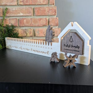 May include: A wooden Christmas countdown calendar with the text "Oh come, oh come Emmanuel" and numbers 1-24. Includes wooden figures of a nativity scene, a star, and a house-shaped sign that reads "Pritzl Family Countdown to Christmas."